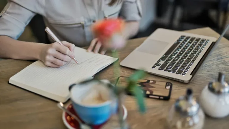 a person writing in a notebook next to a laptop on a desk